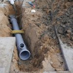 Excavated trench with grey drainage pipe, construction tools, and caution tape for plumbing by Super Plumbers.