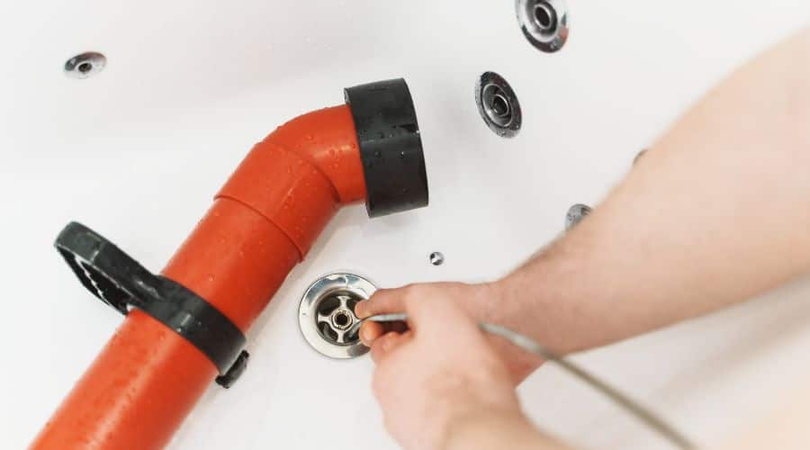 Hand with a flexible drainage snake near a red pipe and metal drain cover, highlighting plumbing repair by Super Plumbers.