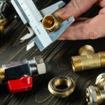 Hand holding a caliper measuring a brass fitting alongside plumbing components at Super Plumbers' workshop in Tyler, TX.